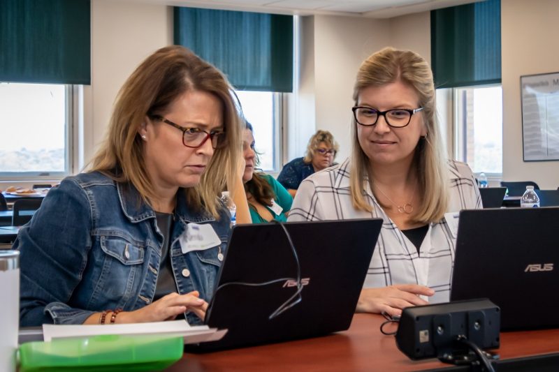 Two women look at a laptop screen sitting on a table in front of them.